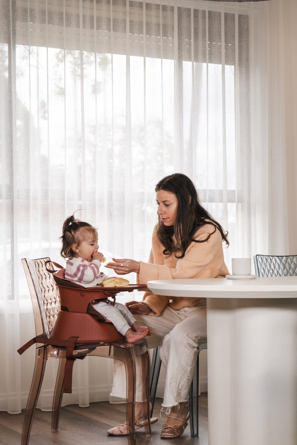 A mother feeding her toddler at a dining table, the child seated in a Hannie in Mahogany Red on a clear dining chair