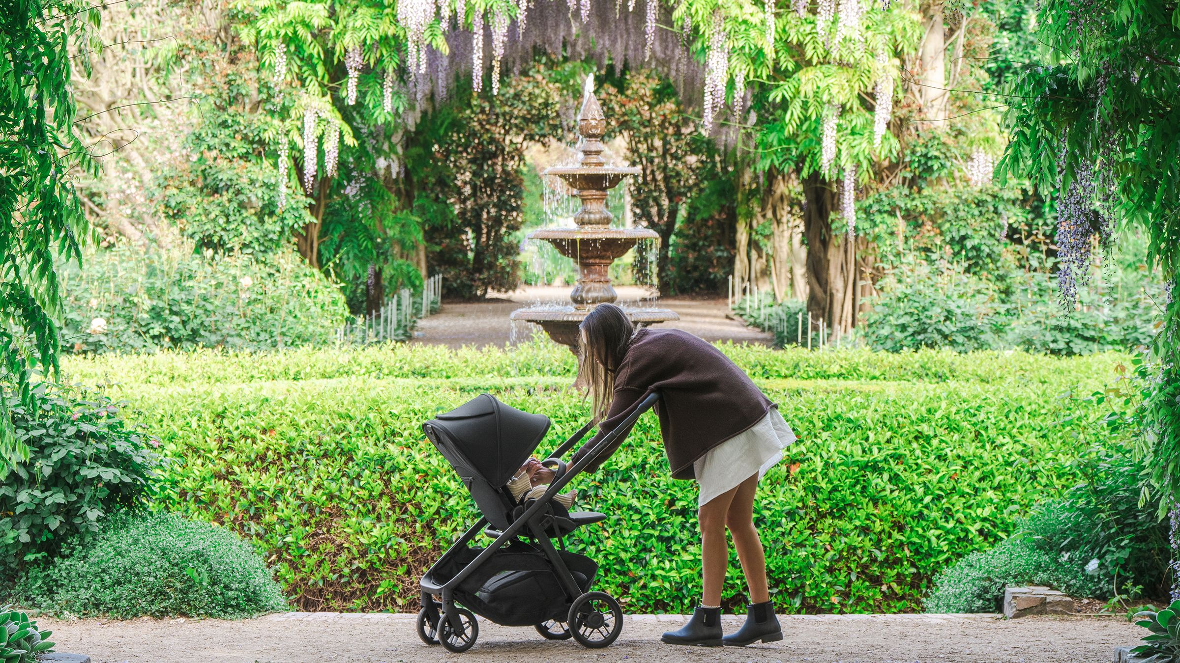 A mum leaning over a Kona pram to her baby, under a wisteria arch with a stone fountain beyond