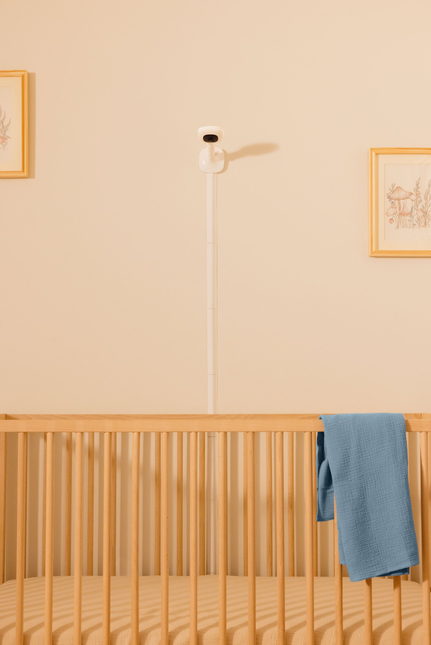 A nursery with a Nanit camera mounted high on the wall above a wooden cot, its cable running neatly down the wall